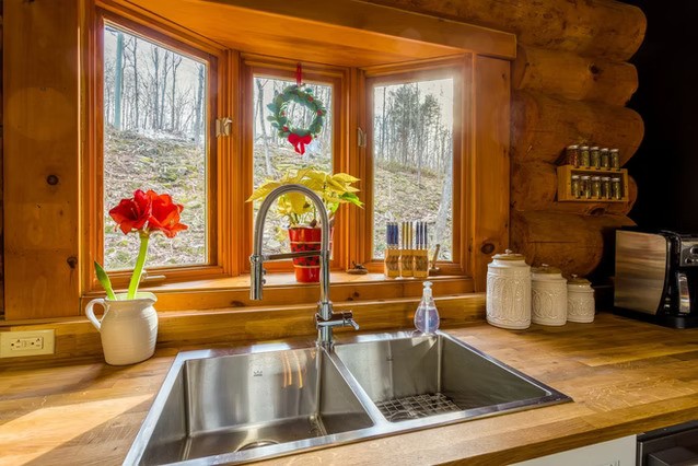 A cozy kitchen sink set in a wooden countertop beneath a window, decorated with plants, a holiday wreath, jars, spice racks, a soap dispenser, and a coffee maker, with a forest view outside.