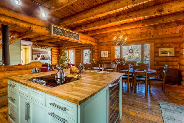 A cozy log cabin kitchen and dining area with wooden walls and ceiling, a center island with a stove, silver kettles, a small decorated Christmas tree, and a dining table by large windows. Warm lighting creates a rustic feel.