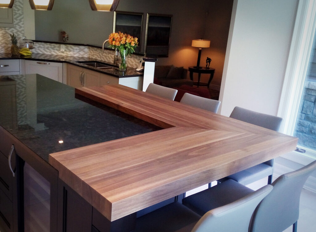 Cuisine moderne avec un grand îlot et une extension de plan de travail en bois en U, entourée de chaises grises. Un vase de fleurs et une corbeille de fruits sont posés sur les plans de travail ; des suspensions sont installées au-dessus.