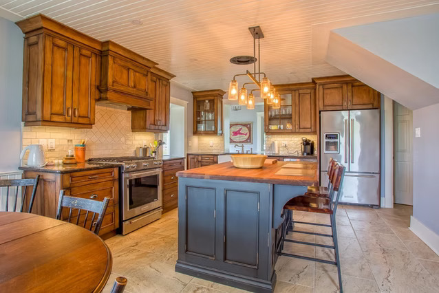 A spacious kitchen with wooden cabinets, a central island with dark blue paneling, hanging pendant lights, stainless steel appliances, and a dining table with chairs on light-colored tile flooring.
