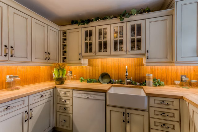 A cozy kitchen with cream cabinets, wooden countertops, a farmhouse sink, warm under-cabinet lighting, and green plants decorating the upper shelves and counter.