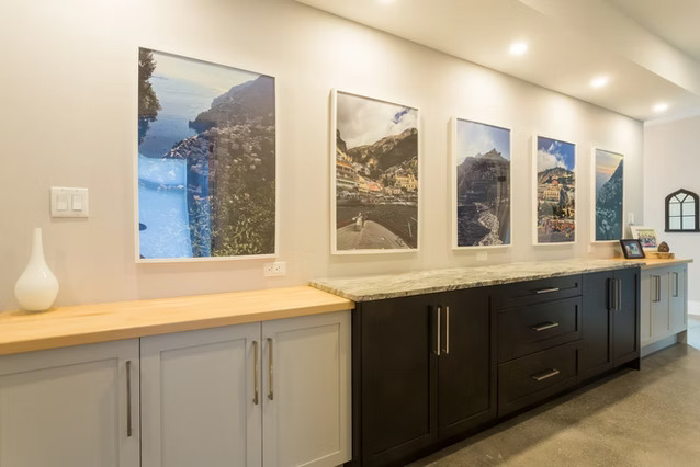 A modern kitchen features light gray and dark cabinets, a wood and marble countertop, and four large framed landscape photos on the wall under recessed lighting.